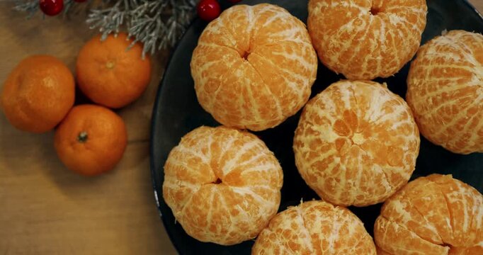 Fresh peeled clementines rest in dark ceramic bowl accompanied by whole citrus fruits and pine needles. Vibrant winter treats evoke warm holiday celebration mood on rustic table surface.
