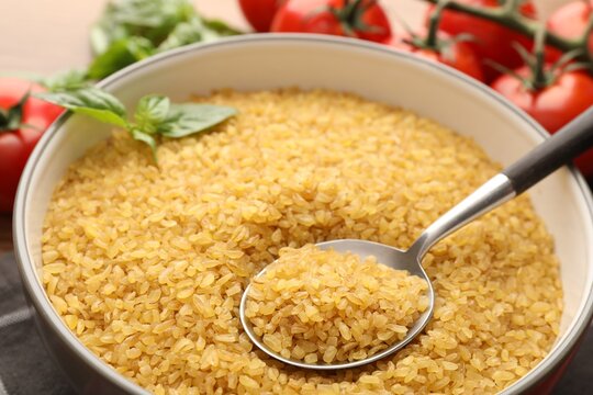 Raw bulgur in bowl, spoon and other products on table, closeup