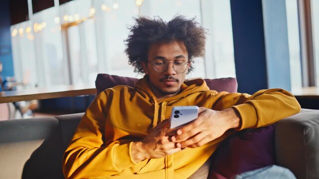Young african american man scrolling phone lounge sofa. Closeup relaxed break.