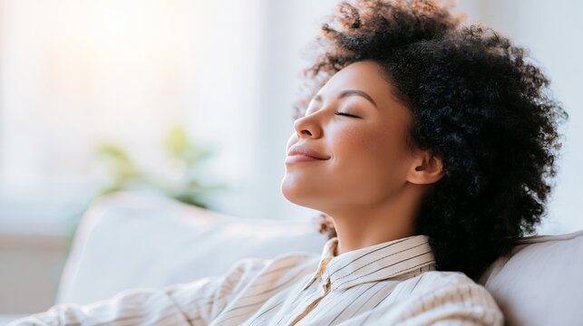 Beautiful woman with natural hair rests peacefully on a soft sofa inside a sunlit room while keeping her eyes closed and smiling gently during a calm moment of quiet reflection