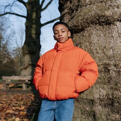 Obraz premium Young boy in bright orange puffer jacket leaning on a tree in an autumn park portrait for seasonal banner and background use