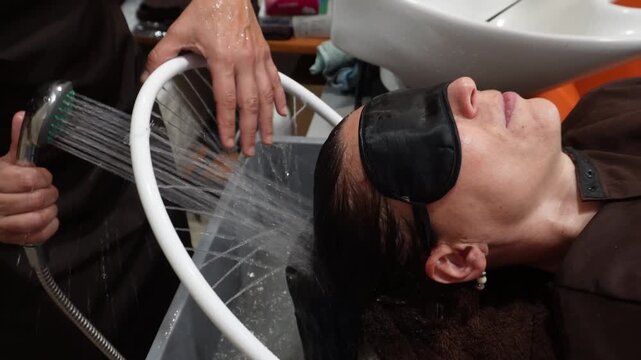 Relaxed woman with an eye mask enjoying a professional head spa massage. A hairdresser gently rinses her hair with a shower jet stream