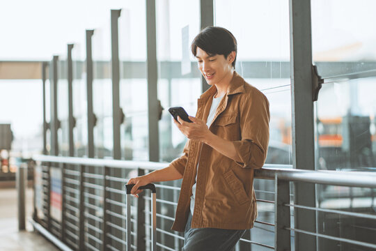 Smiling young man using smartphone with luggage at airport terminal, connected traveler checking mobile phone during journey, air travel, tourism.