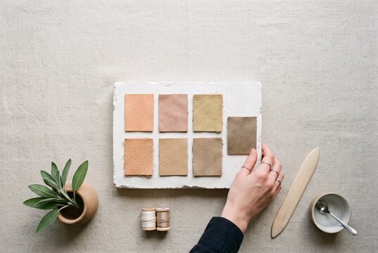 Top view of artisan hand arranging natural leather color swatches on a stone block with crafting tools and plant