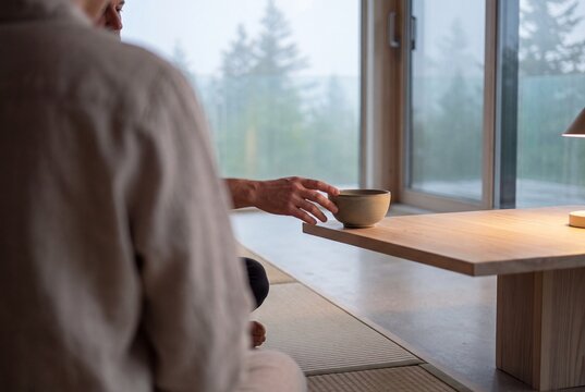 A man sitting on a tatami floor reaches for a ceramic bowl on a low wooden table in a serene minimalist room overlooking a foggy forest