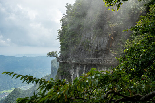 A dramatic view of a narrow, winding skywalk clinging to a sheer rock face, surrounded by dense green vegetation and atmospheric mist in a majestic mountainous region.