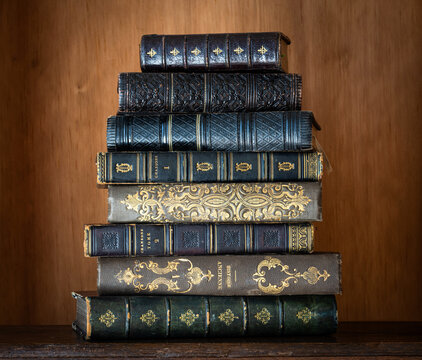 Stack of antique books with richly decorated leather bindings, gilt tooling, raised bands and ornate embossed spines, displayed against a warm wooden background.