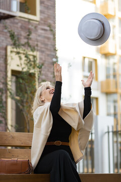 Joyful mature woman stretches toward suspended white hat while sitting on bench. Smiling woman raises both arms during relaxed outdoor play, with cream wrap, black outfit and handbag visible.