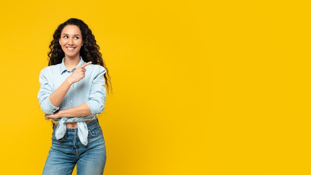 Smiling Young Latina Woman Pointing At Copy Space With Cheerful Expression, Standing In Front Of Yellow Background. Displaying Excitement And Confidence In Business Concept Environment.