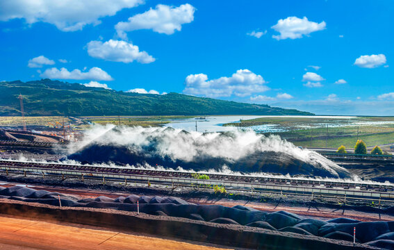 Large iron ore mining operation with conveyor belts and dust suppression water spray over stockpiles against a blue sky.