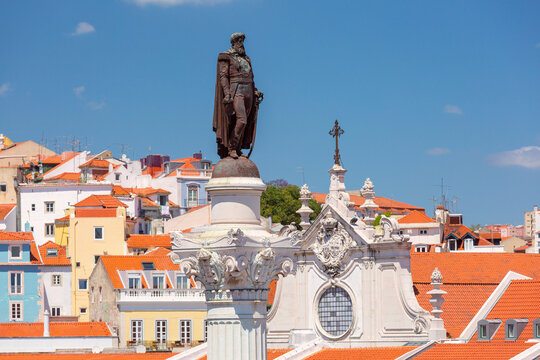 Bronze monument rises above traditional buildings with red tiled roofs in the historic center of Lisbon, Portugal.
