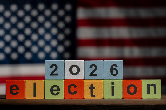US midterms 2026 election concept with wooden blocks, closeup. The word election and numbers 2026 spelled out on colorful wooden toy blocks on wooden table with blurred American flag backdrop