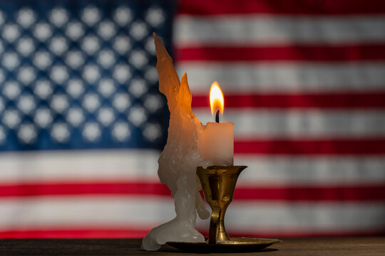 Burning candle against blurred US flag background, closeup. White wax candle with bright flame burns in vintage metal candlestick on wooden table with blurred American flag backdrop