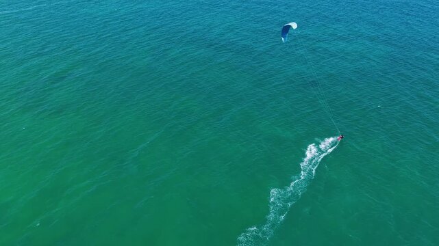 Dynamic kitesurfing action in the ocean. Aerial view of many people riding the waves on the weekend. Colorful parachutes of surfers against the blue sea and sky. Kiteboarding.