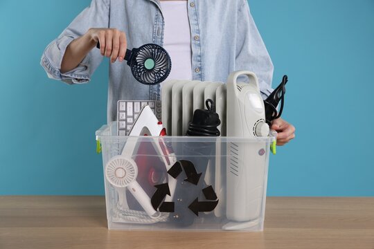 Recycling. Woman putting broken fan into plastic container with different household appliances at wooden table against light blue background, closeup