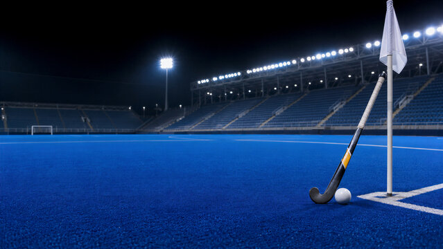 Field hockey stick and ball rest beside a corner flag on an empty blue pitch. Floodlit stadium seating fills the background. World field hockey championships and tournament sport concept.
