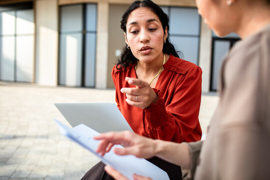 Two businesswomen reviewing documents with laptop in office courtyard