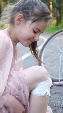 Sad little girl in pain looking at her injured knee with a white bandage after falling off her bicycle, sitting on the road with her pink bike in the background during a summer day