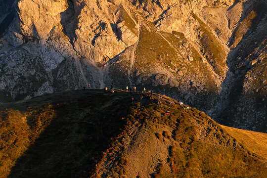 Visitors gather on an alpine ridge at Passo Giau to watch the sunset, surrounded by warm golden light, grassy slopes and rugged Dolomite rock walls in northern Italy.