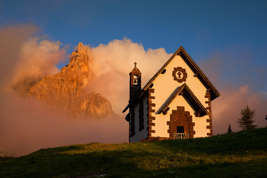 Chiesetta della Madonna Assunta at Passo Rolle illuminated by warm golden light with Cimon della Pala rising through clouds in the Pale di San Martino, Italian Dolomites. Colors: orange, green, blue.