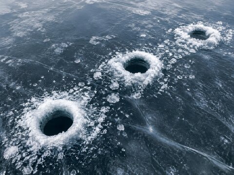 Three drilled ice fishing holes cut in a line across dark ice with slush rings and shaved ice