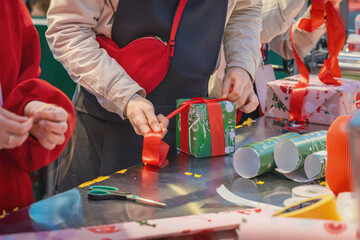 Obraz premium Hands Making Bow on Wrapped Christmas Present. Person finishes wrapping green Christmas box present with wide red ribbon at festive work table. Close-up handmade gift preparation, holiday packaging