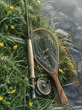 Fly fishing rod and wooden landing net resting on a grassy bank above a clear stream
