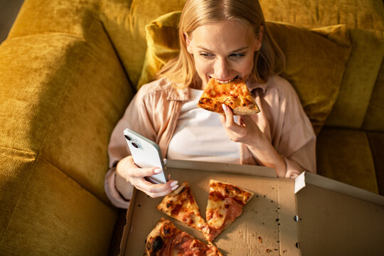 Young woman eating pizza while using smartphone at home