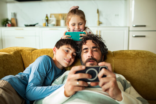 Father and children playing mobile games on home sofa