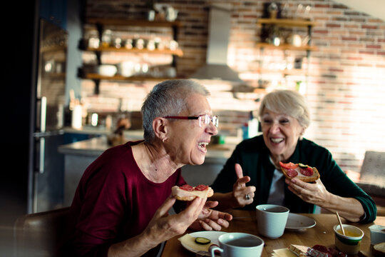 Two senior women laughing while eating breakfast at home kitchen