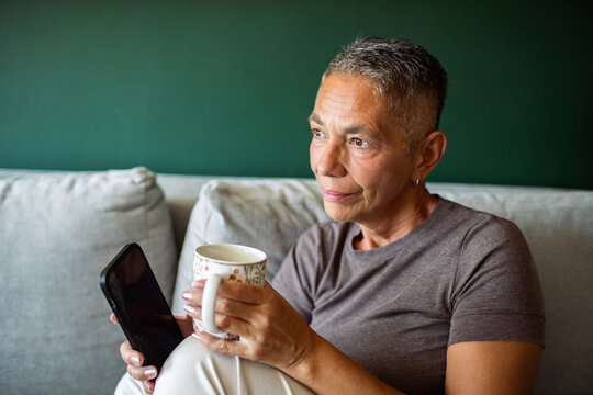 Senior woman relaxing on sofa with coffee and smartphone at home
