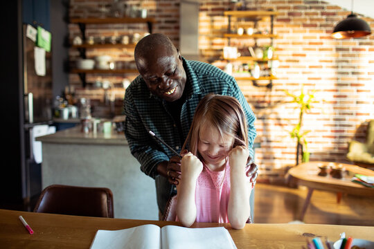 Smiling man helping young girl with homework at kitchen table at home