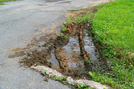 Muddy Road Edge with Deep Tire Tracks, Water Puddles, and Discarded Grass Clippings on a Cracked Asphalt Surface Bordering a Lush Green Lawn