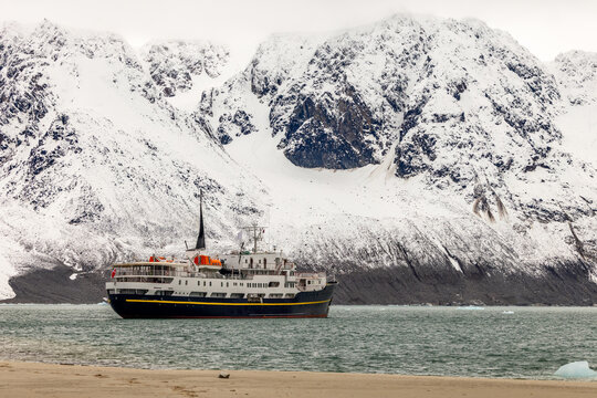 Passenger expedition ship sailing through Arctic water beneath massive snow-covered mountains, Spitsbergen, Svalbard, Norway