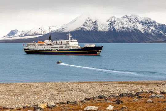 Passenger expedition ship anchored in Arctic fjord with small zodiac boat, snowy mountain range and remote summer coastline, Spitsbergen, Svalbard, Norway