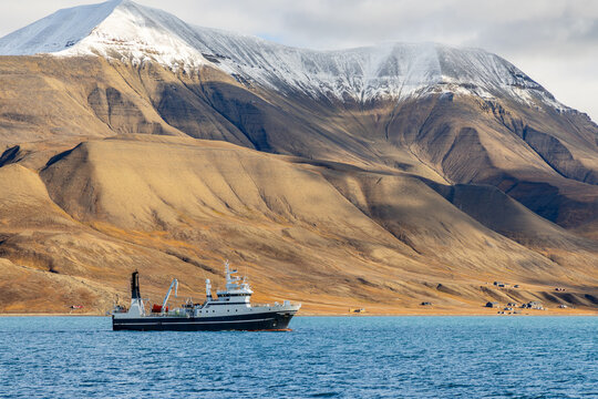 Ship navigating a remote fjord beneath monumental tundra mountains with snow on the ridges and scattered coastal buildings, Spitsbergen, Svalbard, Norway