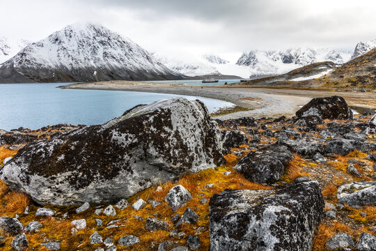 Rugged Arctic summer landscape with massive lichen-covered boulders, calm fjord water, snowy mountains and distant expedition ship, Spitsbergen, Svalbard, Norway