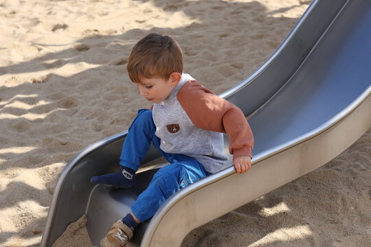 Little boy sliding down a playground slide in a sandy park on a sunny day. The candid scene captures childhood joy, outdoor activity, and playful moments in a safe environment.