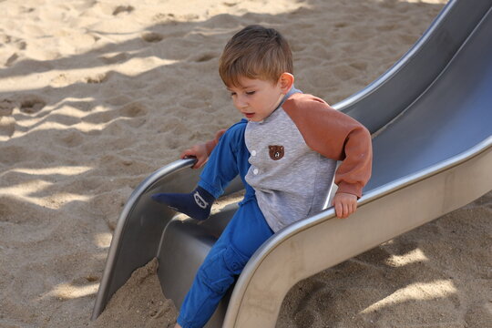 Little boy sliding down a playground slide in a sandy park on a sunny day. The candid scene captures childhood joy, outdoor activity, and playful moments in a safe environment.