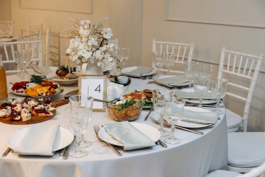 Festive round banquet table setting with table number four, food platters, floral centerpiece, and white chiavari chairs.
