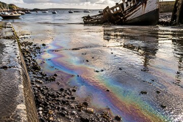Obraz premium Iridescent diesel film spreading across a wet concrete boatyard slipway beside wreckage at low tide