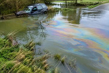Obraz premium Iridescent diesel film spreading across floodwater in a flooded ford beside a stranded wreck in a river meadow