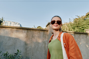 A confident woman stands outdoors in a green dress and orange cardigan, smiling at the camera. Sunglasses shade her eyes as she poses against a weathered wall under bright daylight, conveying casual © SHOTPRIME STUDIO