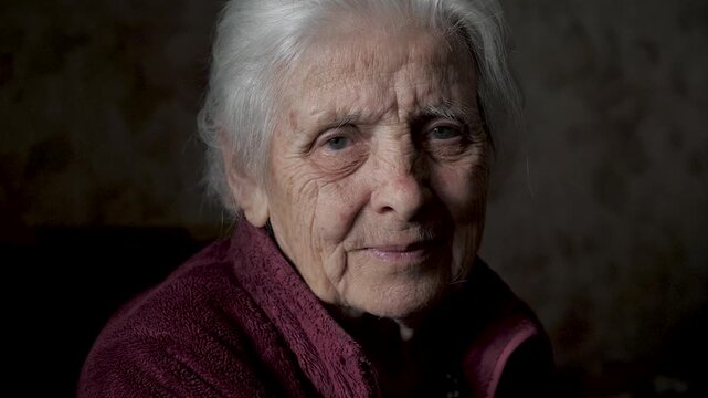 Elderly woman with deep wrinkles and gray hair sitting in dark room looking at camera with gentle and serene smile. Old female conveying wisdom and lifetime of experience through her eyes