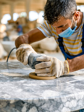 A worker in a face mask and protective gloves uses an electric orbital sander to polish the smooth surface of a round grey marble table in a workshop.