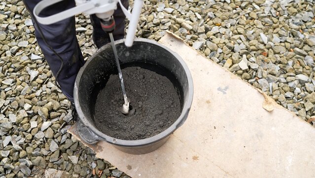 Construction worker mixing concrete mortar with an electric drill mixer in a bucket on a gravel surface