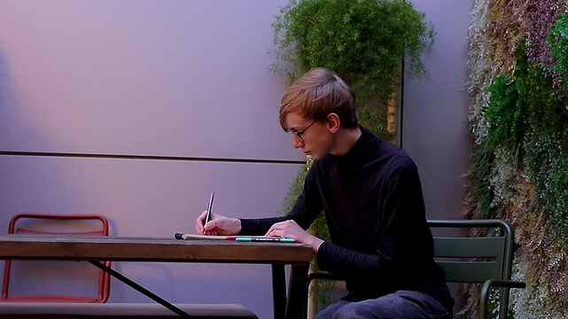 Young man wearing a black turtleneck and glasses passionately drawing or making notes with a pen in a sketchbook while sitting in a patio with green plants.