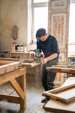 Professional handicraftsman cutting part of wood from antique window frame using handheld wood router, man woodworker processing and rebuilding vintage wooden frame with electric plunge router machine