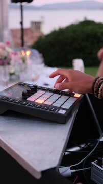 Close-up of a musician's hand playing a drum machine, creating electronic music beats for a celebration event by the lake during a beautiful twilight with soft, ambient lighting. Vertical video