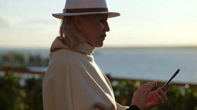 Woman studies smartphone beside calm coast in evening light. Elegant woman wears wide brim hat and light coat, holding phone with focused calm while standing near water in coastal lifestyle setting.